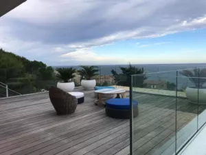 Terrasse en bois avec assises extérieures et garde-corps en verre devant la mer.