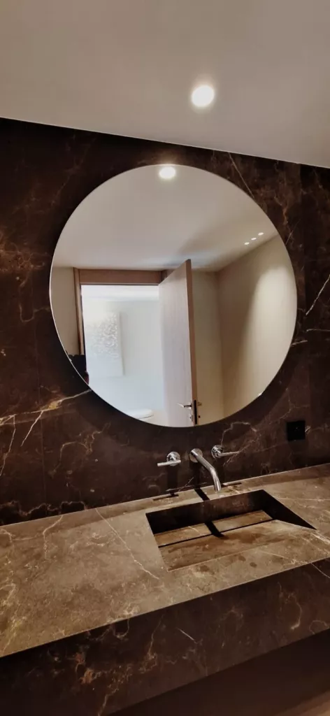 Grand miroir rond au-dessus d’une vasque et devant un mur en pierre sombre.