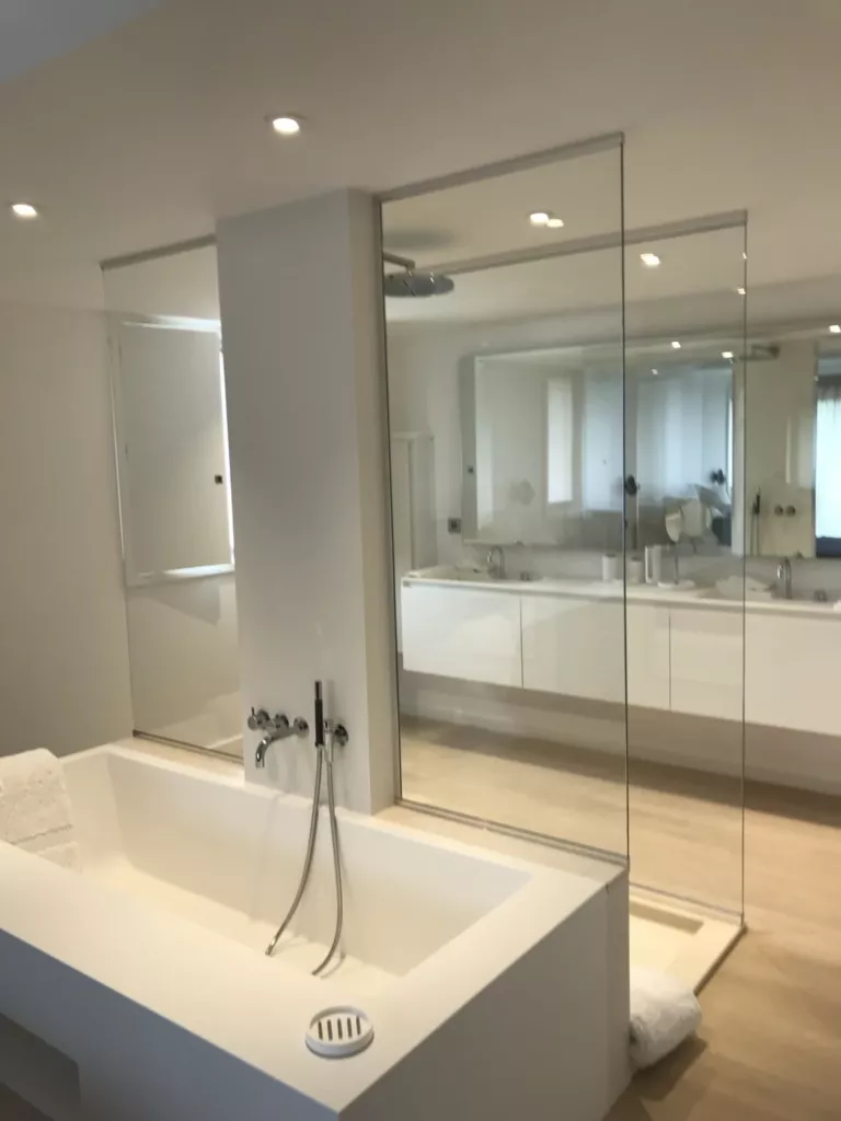Salle de bains claire avec baignoire, grands miroirs et parois vitrées transparentes.