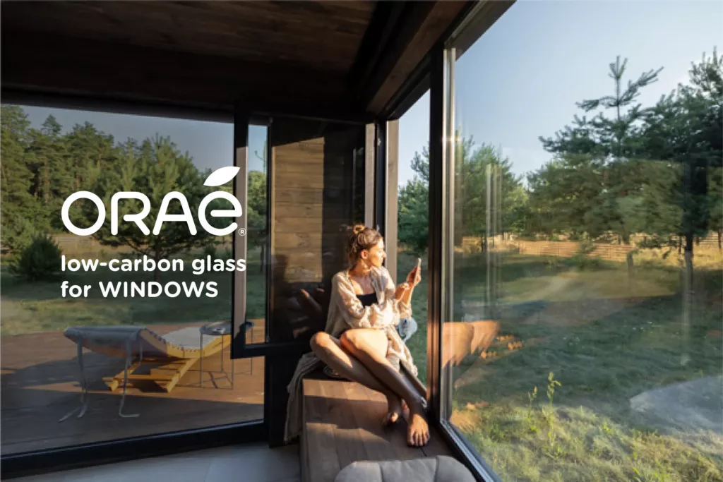 Séjour baigné de lumière derrière de grandes baies vitrées, avec la mention « ORAÉ low-carbon glass for windows » (illustration Saint-Gobain Glass, ORAÉ)